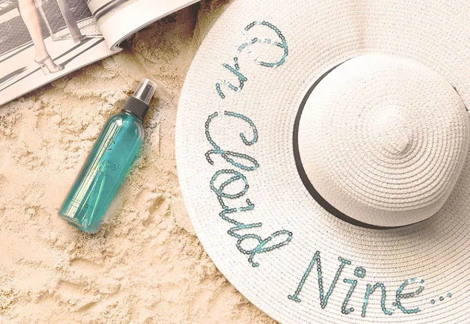 Image of a white hat embroidered with 'On Cloud Nine' in blue sequins next to a bottle of Magical Potion on the sand.