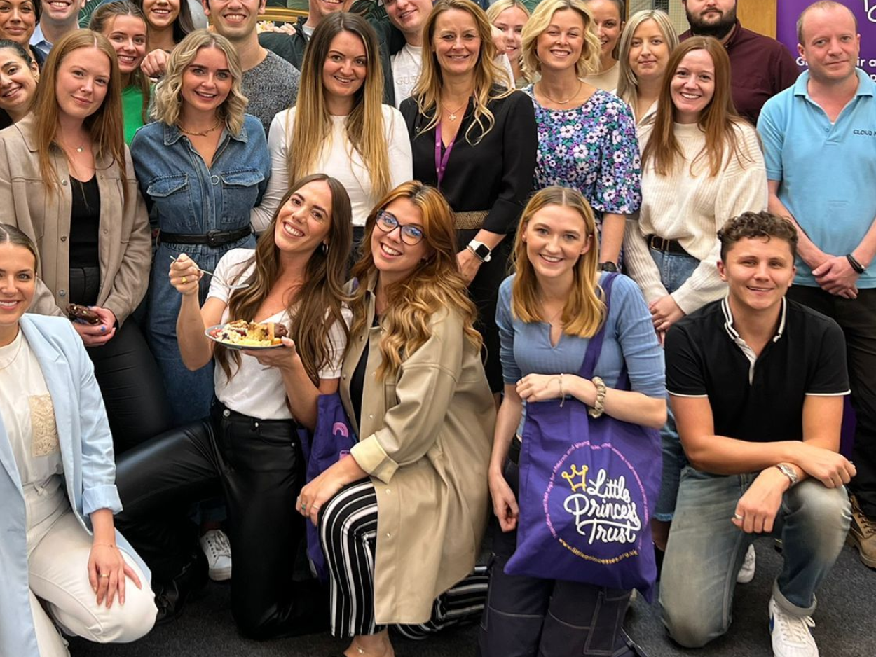 The Little Princess Trust team smiling in a group. One woman is eating baked goods that have been made to raise money for the charity.