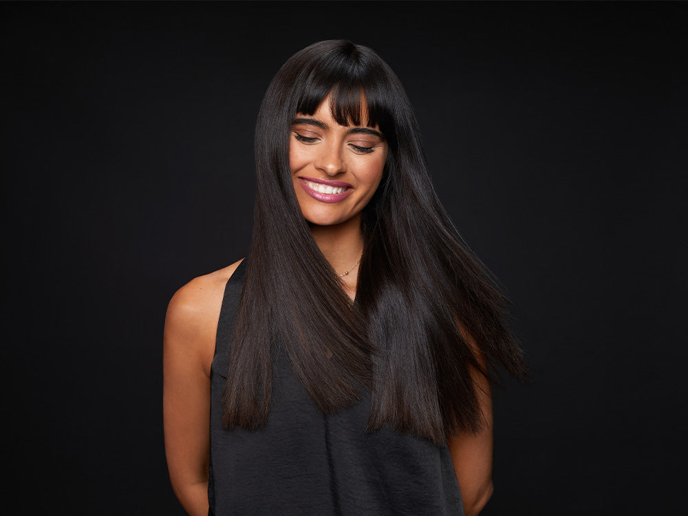 Model with long, straight dark hair smiling and looking towards the ground.
