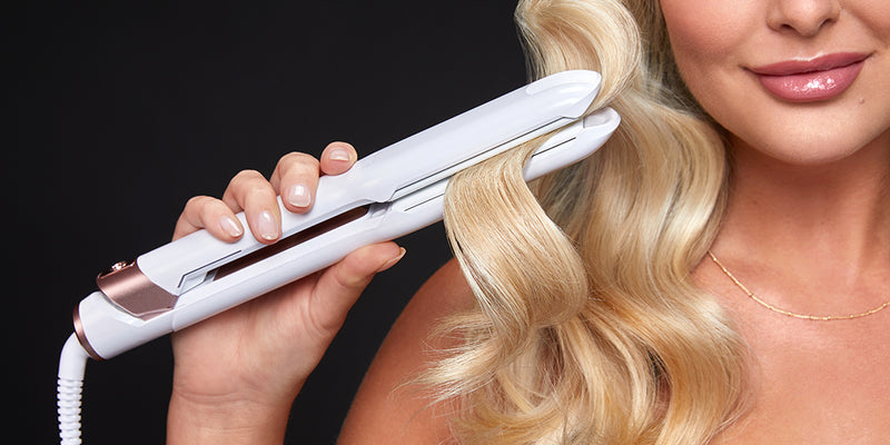Blonde model smiling while running a white hair straightener through waved hair.