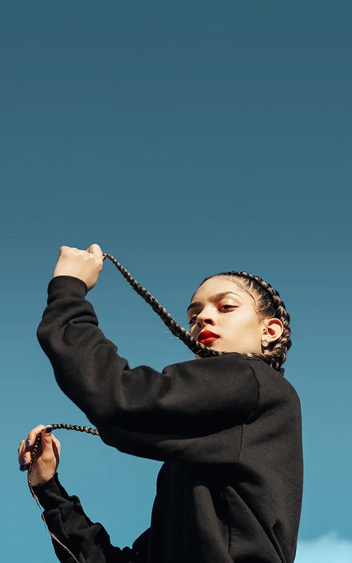 A woman turned to the side and holding her long black braids in front of a blue sky.