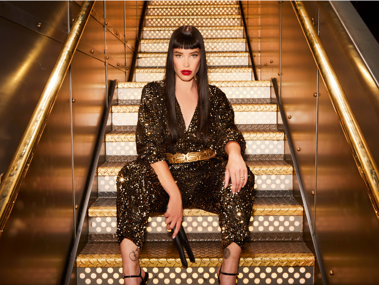 A black haired woman dressed in a sequin jumpsuit sitting on gold and silver stairs holding a pair of CLOUD NINE hair straighteners.