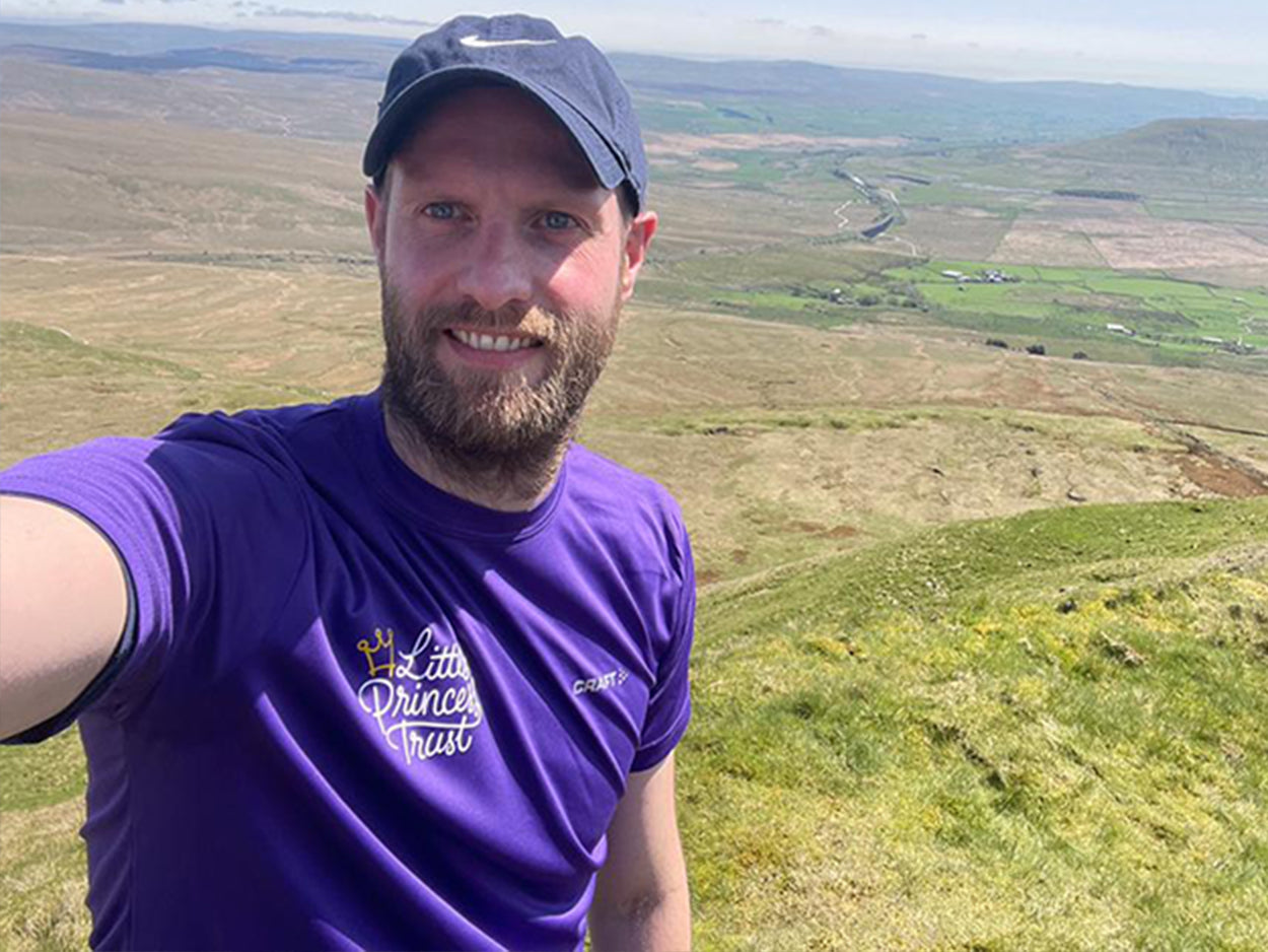 A man completing a charity walk for Little Princess Trust. He is stood on top of a hill wearing a Little Princess Trust t-shirt.