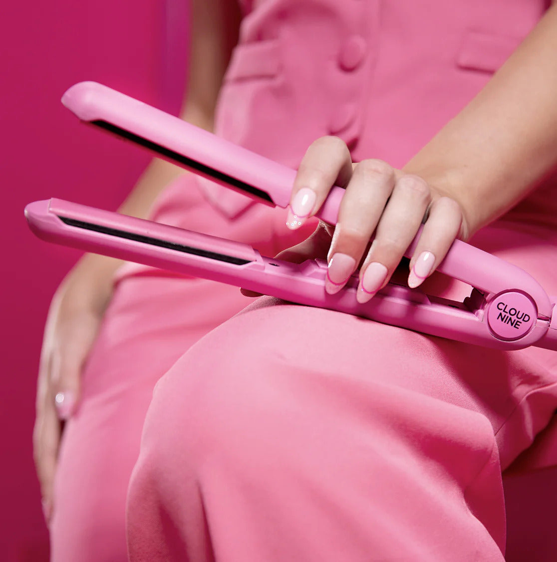 Model in all pink outfit holding a pink CLOUD NINE hair straightener on knee.