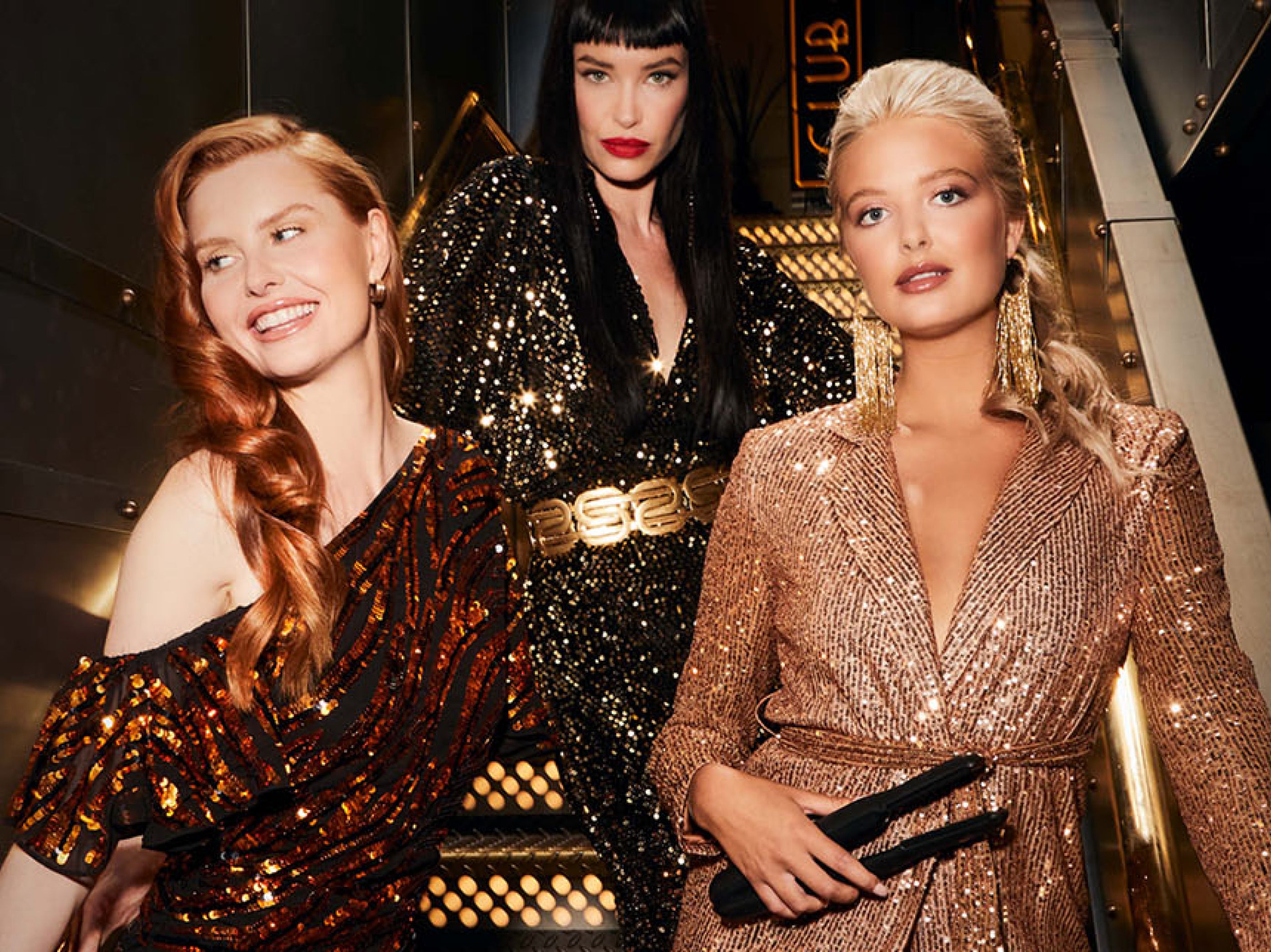 3 models, one with red hair, another with black hair and one with blonde hair, all wearing glittery outfits. The blonde model is holding The Cordless Iron.