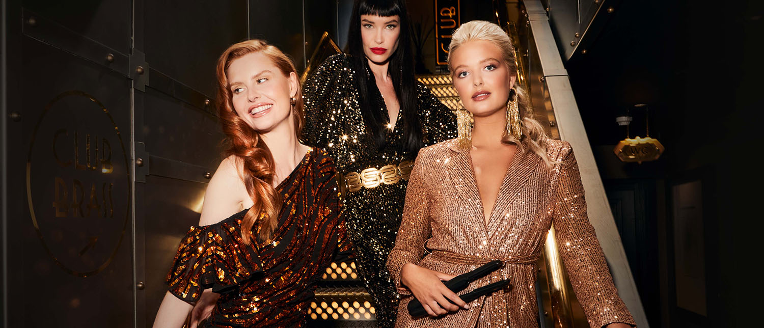 Three women wearing sequin outfits standing on some stairs. A smiling red haired woman, a pouting black haired woman and a blonde haired woman holding a CLOUD NINE straightener.